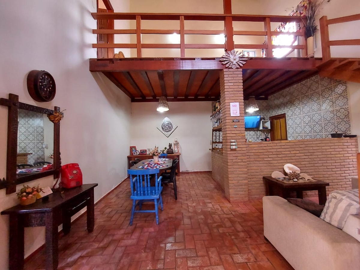 The interior space features an open layout with a high ceiling, showcasing a mix of rustic and cozy elements. A dining area is seen with colorful chairs, while a bar area is positioned along one wall. Natural light enters through a window, enhancing the inviting atmosphere.