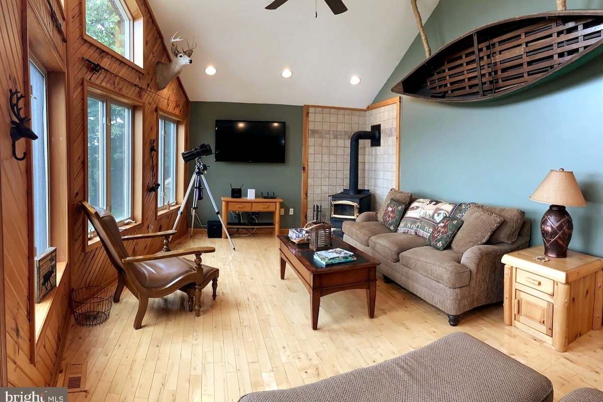 A spacious living room is characterized by wooden walls and a light wooden floor. Large windows flood the area with natural light. A comfortable couch and a recliner provide seating, while a wood-burning stove adds warmth. A telescope and a mounted canoe are also present.