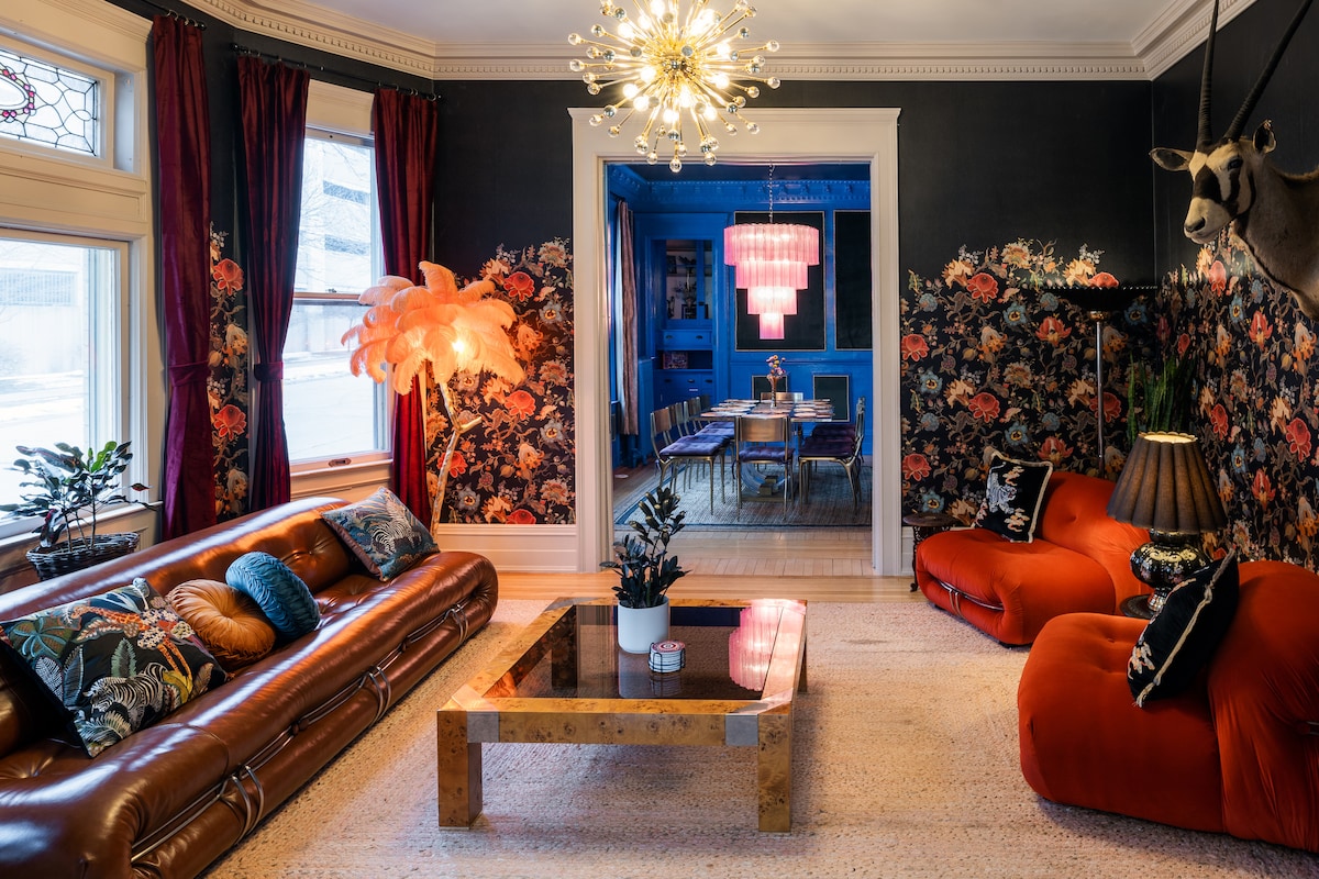 A stylish living room features a combination of modern and vintage decor. A leather sofa and two vibrant red chairs are arranged around a wooden coffee table. Decorative plants and unique light fixtures add character, while floral wallpaper and a colorful dining area can be seen in the background.