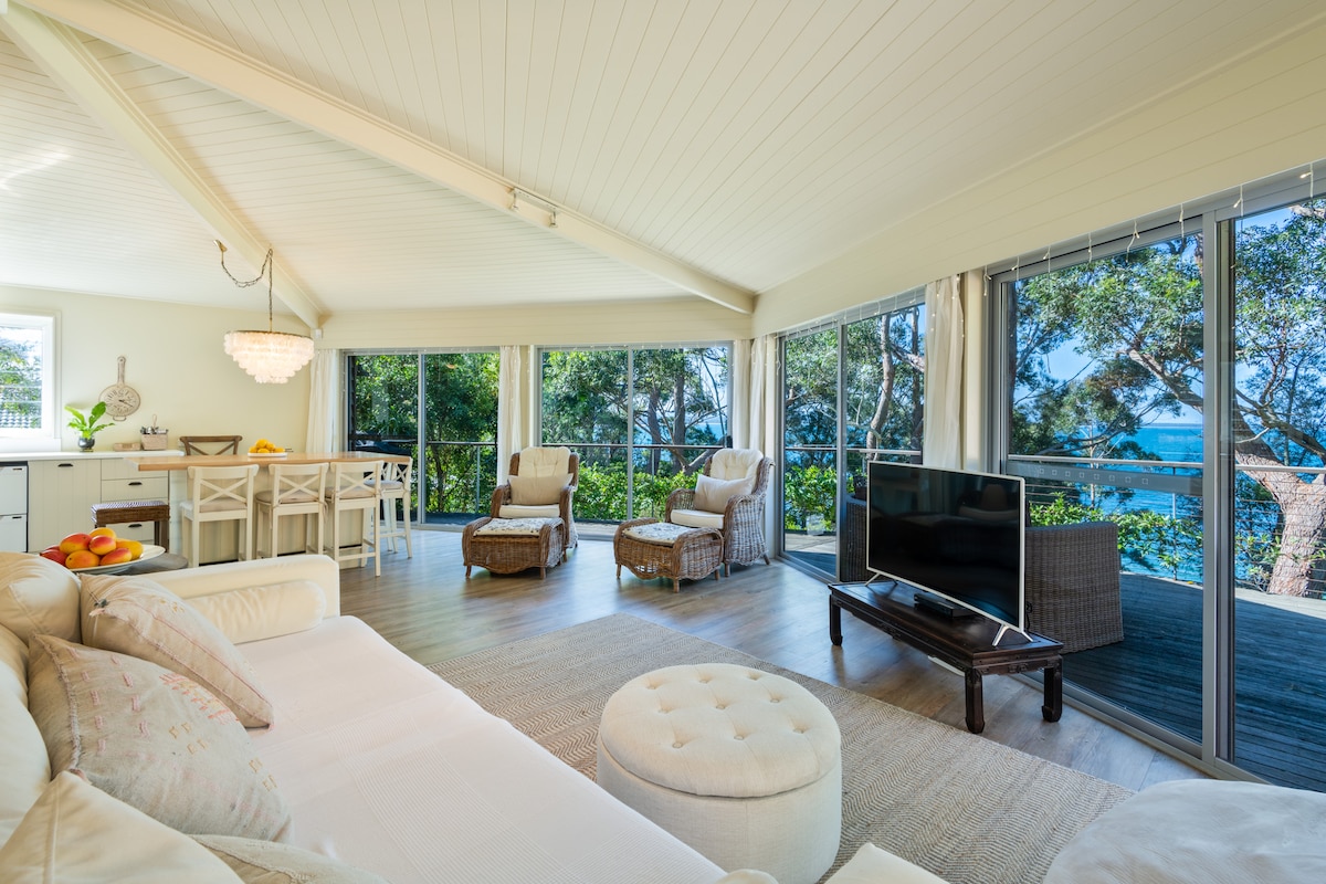 A bright living area features large glass doors that open to a view of the trees and waterfront. Comfortable seating includes a spacious white sofa and armchairs in a natural woven texture. The room is complemented by light wood paneling and a soft area rug.