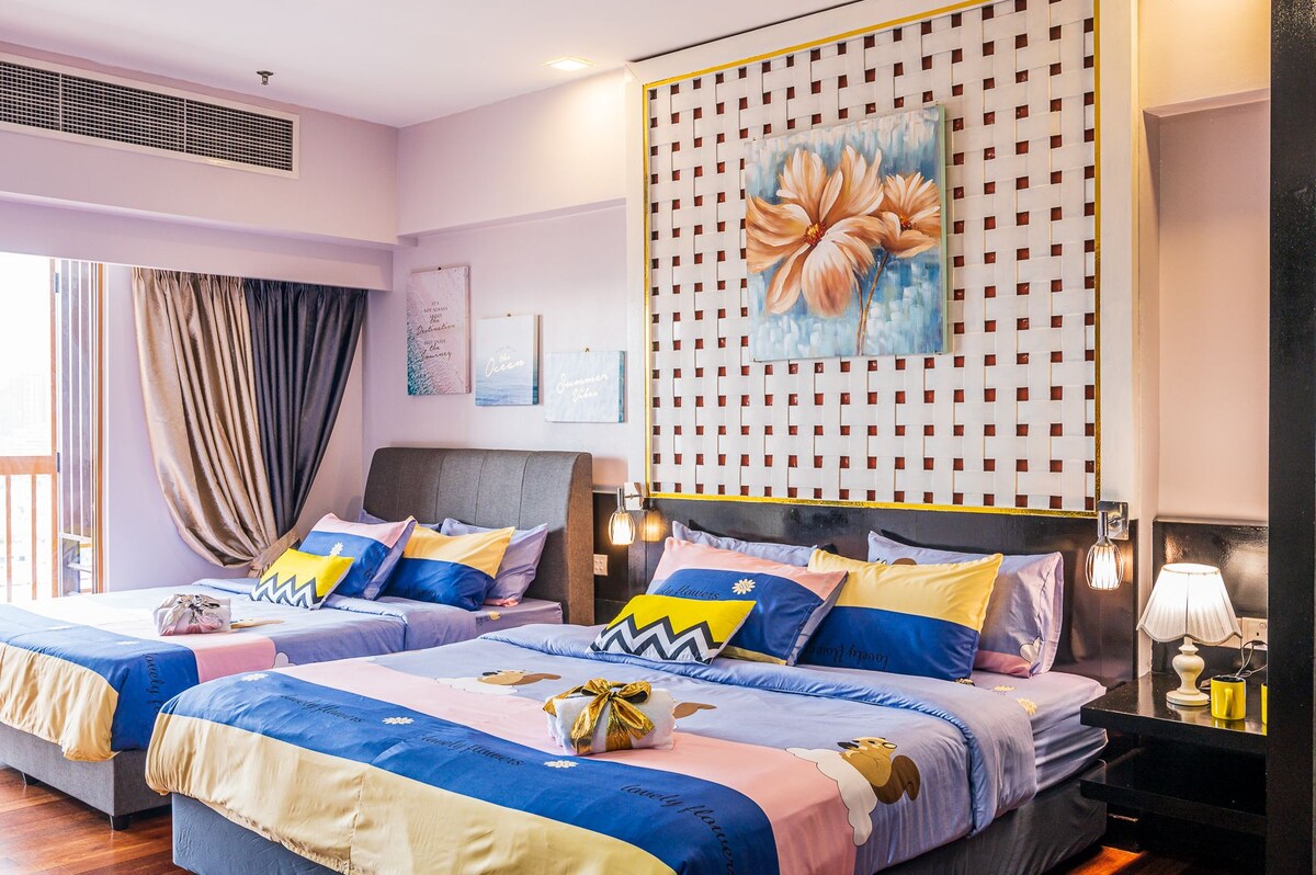 A spacious sleeping area is balanced with two double beds dressed in colorful linens and decorative pillows. The wall features a large, floral art piece framed by a grid design. Soft lighting is provided by bedside lamps, and natural light enters through the window.