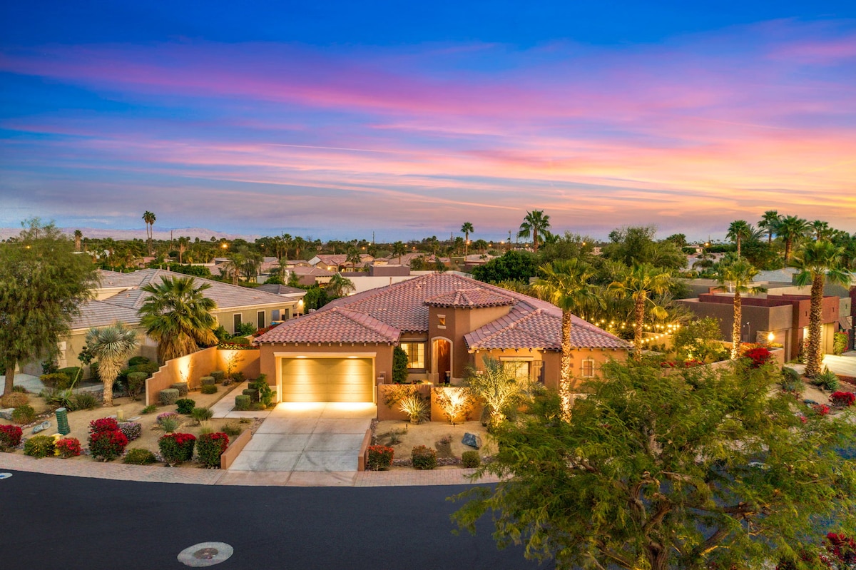 The exterior of a single-story residence is captured during a colorful sunset. The home features a terracotta tiled roof and is surrounded by lush landscaping, including palm trees and vibrant vegetation. A driveway leads to a two-car garage, with outdoor lighting enhancing the welcoming ambiance.