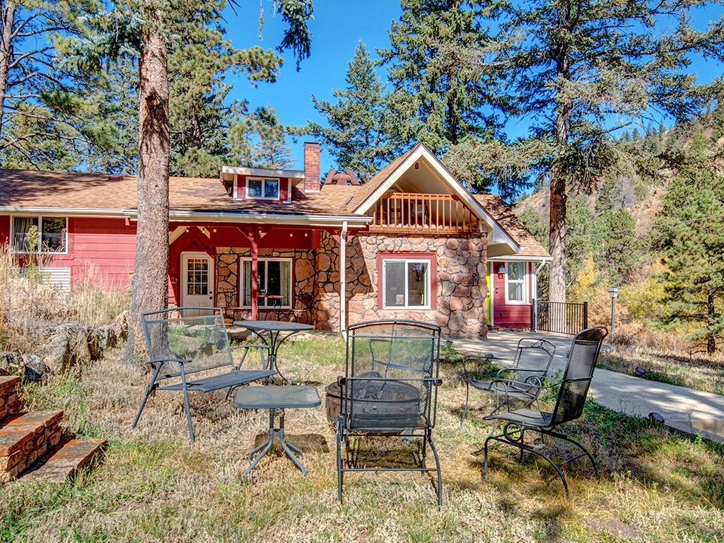 The exterior of the cabin is surrounded by tall pine trees, featuring a combination of stone and wooden siding. A seating area with multiple metal chairs is visible on the grassy lawn, providing a relaxed outdoor space for gathering and enjoying the natural surroundings.