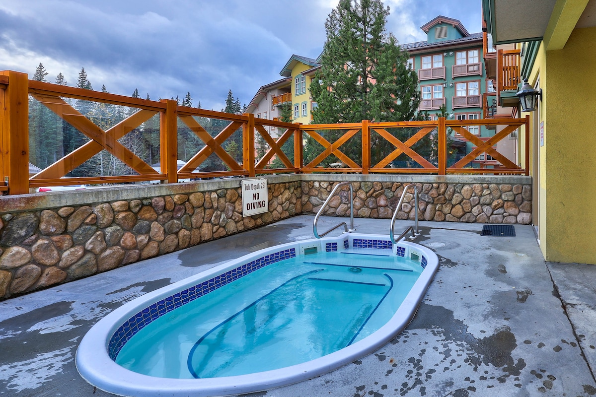 A shared hot tub is situated on a patio, surrounded by a wooden fence with a natural stone wall. The water is clear, and the area features a concrete surface. Scenic views of trees and a building can be seen in the background.