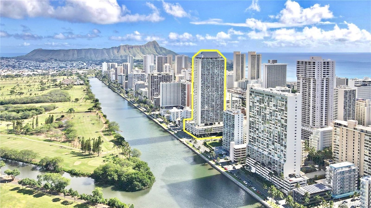 The studio apartment building is prominently positioned amidst the vibrant landscape of Waikiki. Surrounded by a mix of modern high-rises and lush greenery, the scene highlights the nearby coastline, showcasing a blend of urban and natural beauty.