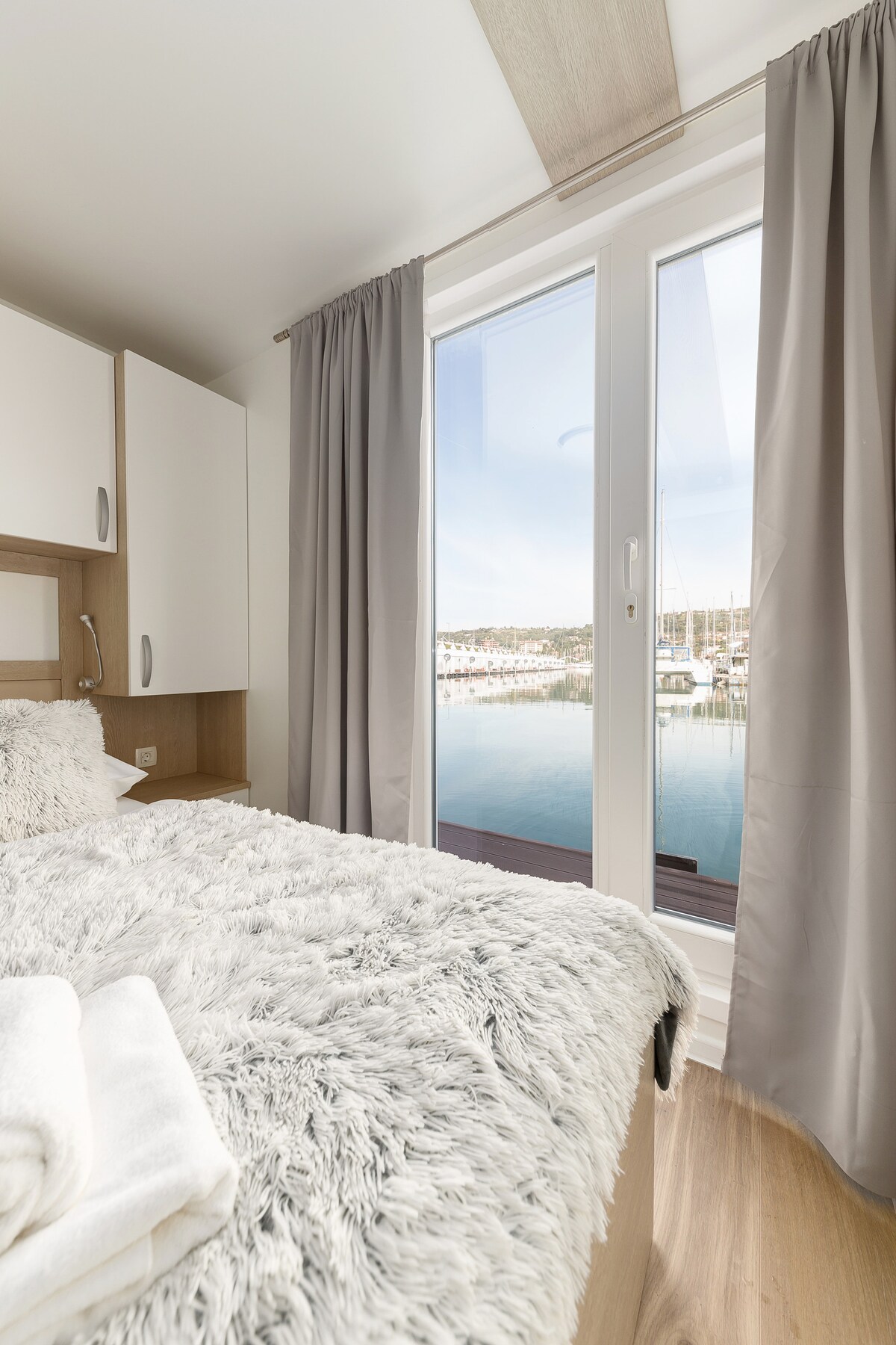 A bright bedroom features a plush, textured blanket draped over a spacious bed. Light enters through large windows adorned with soft gray curtains, framing scenic waterfront views. White towels are neatly arranged nearby, contributing to a fresh and inviting atmosphere.