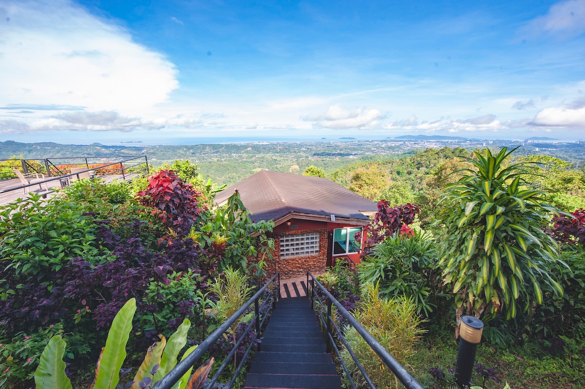 A hillside cottage is framed by lush greenery, showcasing vibrant plants and flowers. The view extends across the valley, revealing the South China Sea and the distant cityscape under a clear blue sky. A flight of steps leads down towards the cottage entrance.