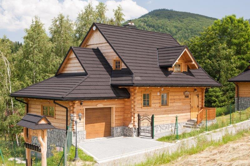 A wooden cabin designed with natural materials is set against a backdrop of lush greenery and mountains. The structure features a gabled roof and a spacious driveway leading to a garage. Fencing adds privacy to the inviting exterior space.