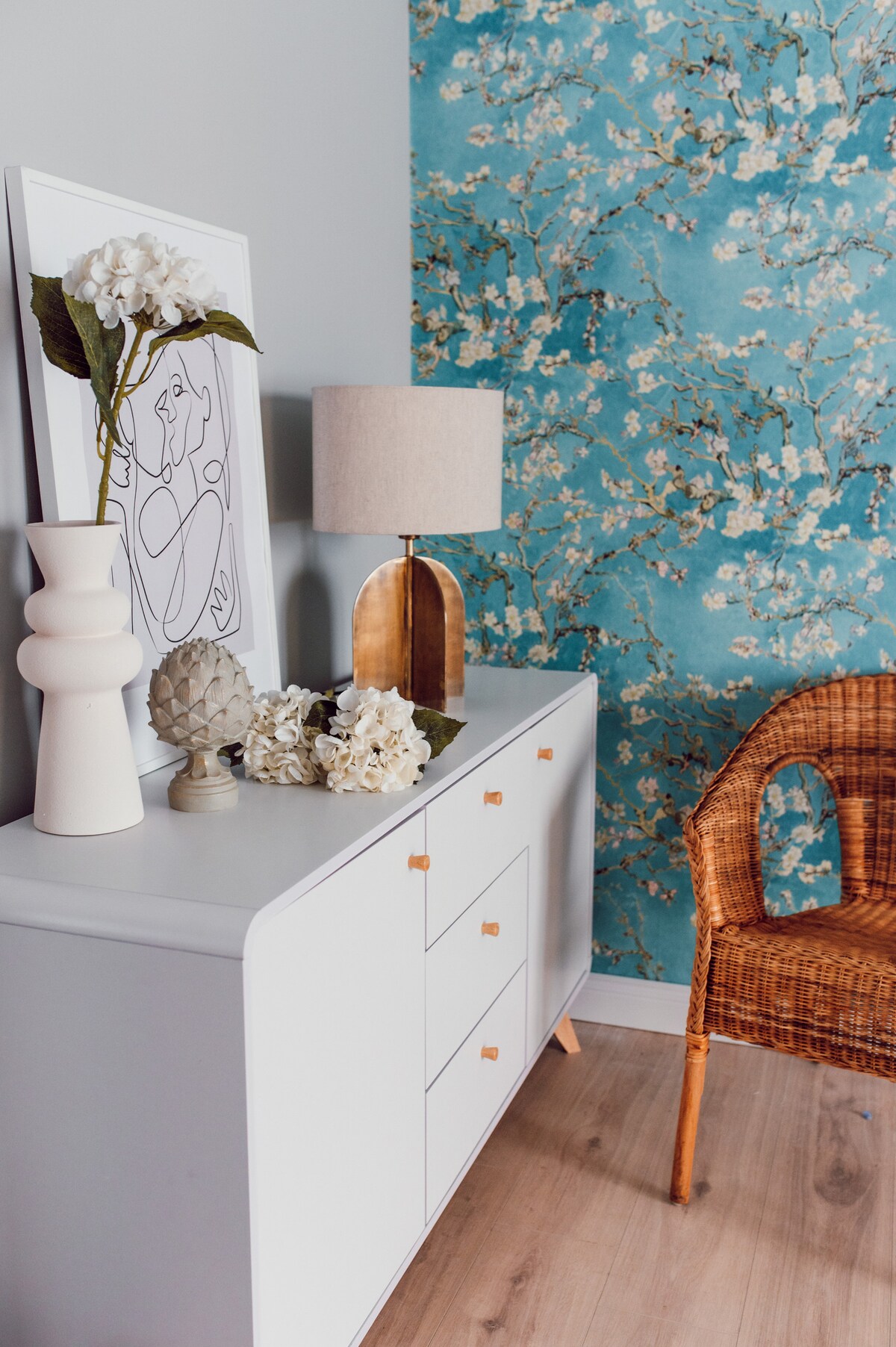 A light-colored dresser is accompanied by a decorative lamp and various floral arrangements. A piece of artwork is displayed alongside a patterned turquoise wall featuring floral designs. A woven chair adds a touch of texture to the elegantly styled space.