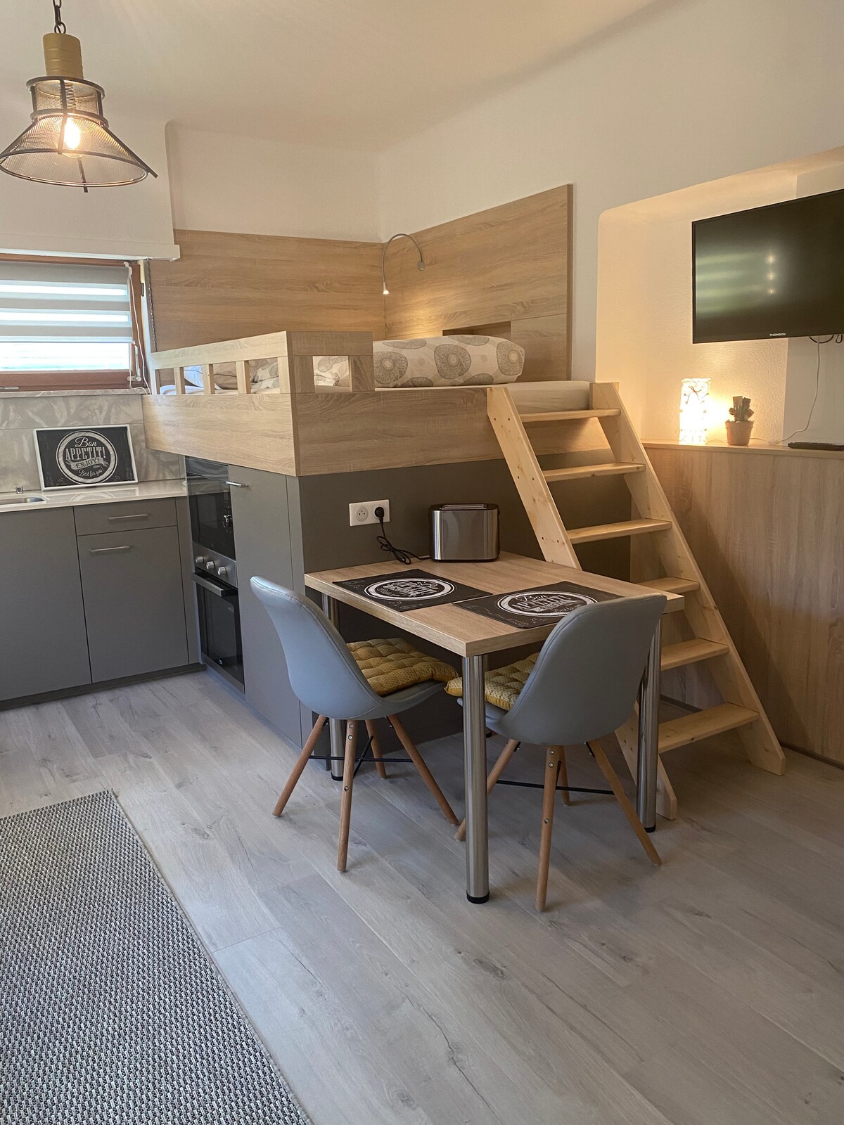 A modern kitchen area with wooden accents features a table set for two. Grey chairs are positioned around the table, and a small staircase leads to a lofted sleeping area. Appliances such as a kettle and a stove are visible, along with a television mounted on the wall.