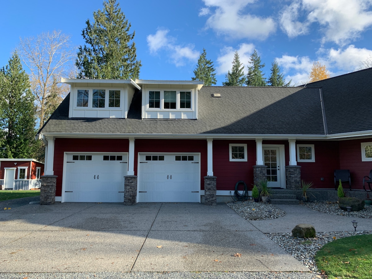 The front of a two-story home is showcased, featuring a red exterior with white trim and a spacious driveway. Two garage doors are visible beneath a stone accent arch, while large windows above the garage provide natural light. Surrounding trees contribute to a serene atmosphere.