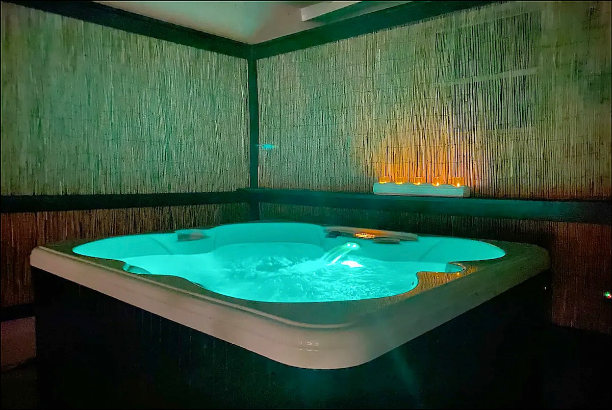 A well-lit hot tub is situated within a private enclosure, featuring soothing turquoise water and ambient lighting. The surrounding walls are adorned with natural materials, providing a serene and intimate atmosphere for relaxation.
