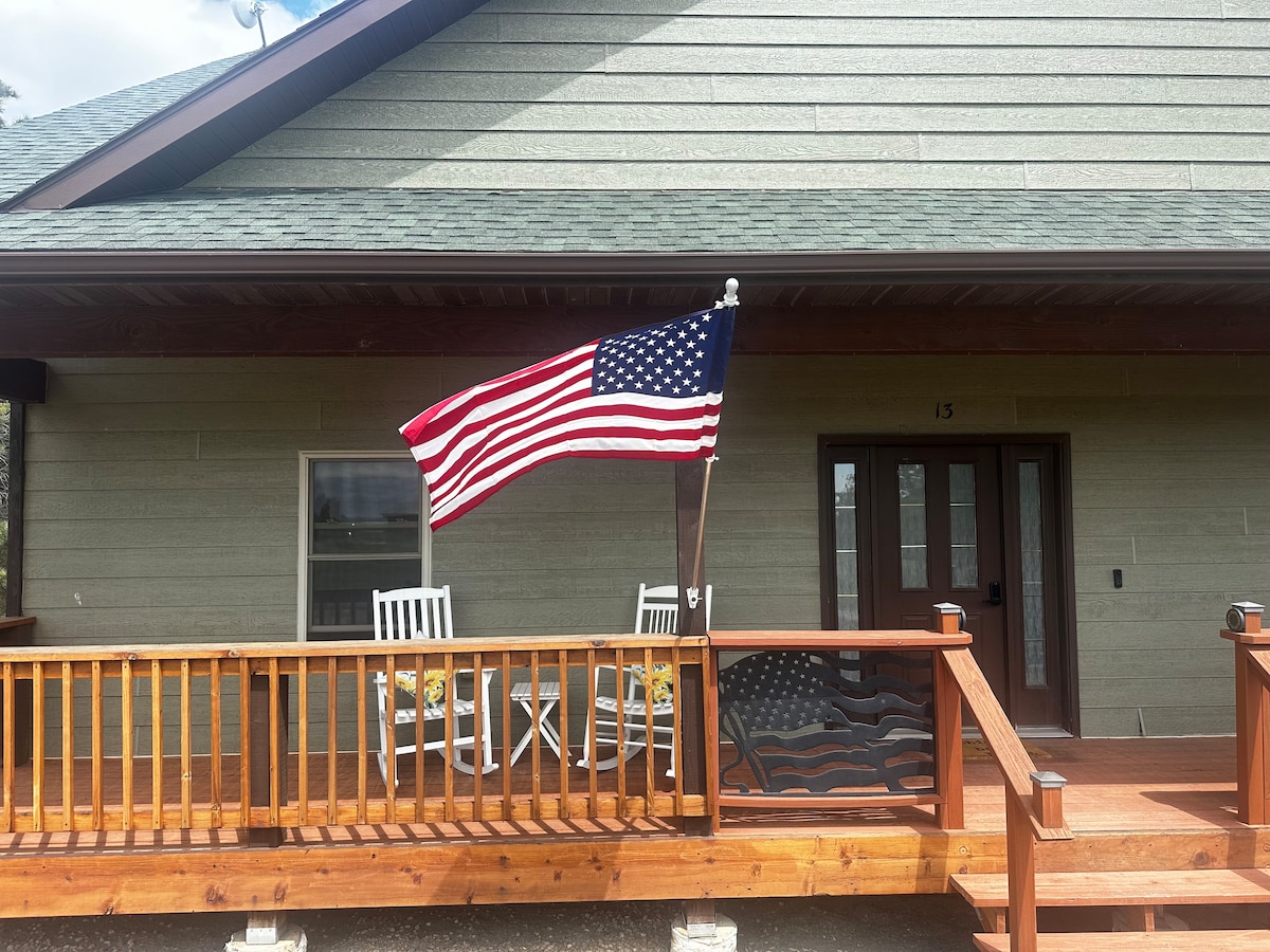 A front porch is visible, featuring a wooden railing and two white chairs positioned beside a table. An American flag is displayed prominently, waving gently in the breeze. The entrance to the home is framed by double doors, leading into the interior.