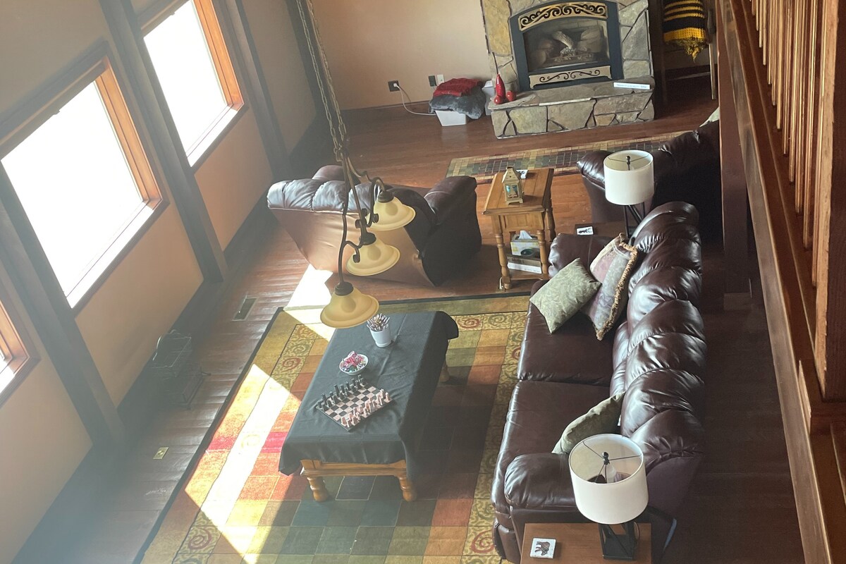 A spacious living area features a brown leather sofa and loveseat arranged around a coffee table. A decorative area rug adds color to the wooden floor. Natural light floods the space through large windows, illuminating a stone fireplace at the far end.