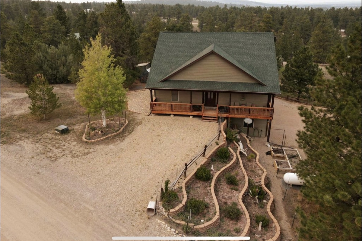 An aerial view of a well-maintained house set in a natural environment, surrounded by trees. A landscaped yard is visible, featuring winding paths and neatly arranged flower beds. A spacious porch leads to the entrance of the home, providing an inviting outdoor space.