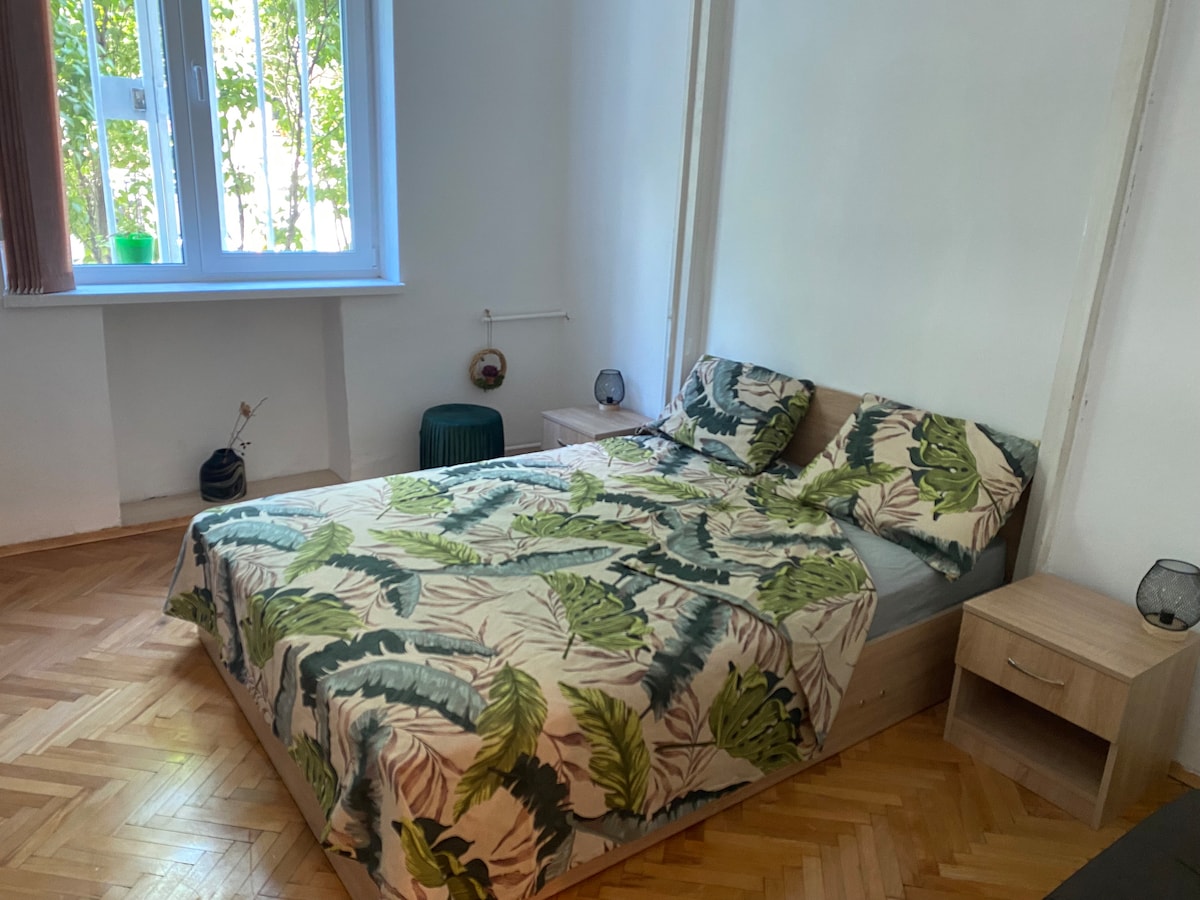 A bright bedroom is depicted, featuring a bed adorned with a leafy green patterned duvet cover. Natural light filters through the window, illuminating the wooden floor. A small side table with lamps is positioned next to the bed, contributing to the room's functional layout.