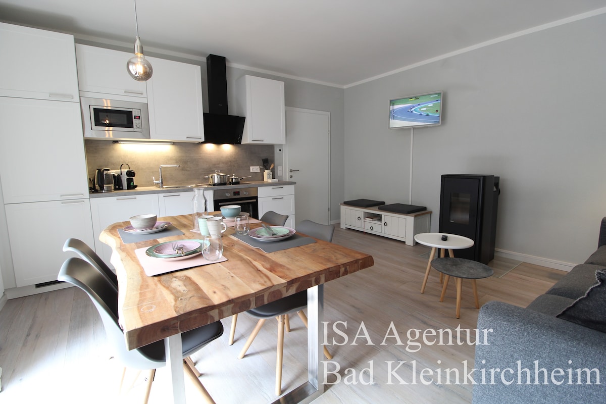 A modern kitchen area features sleek white cabinets, stainless steel appliances, and a wooden dining table set for four. A cozy living space includes a pellet stove and a comfortable sofa, with light colors enhancing the overall design. A television is mounted on the wall.