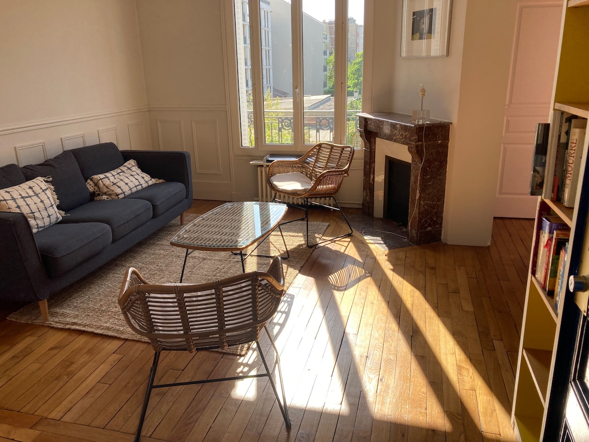 A bright living room features a dark sofa and two rattan chairs flanking a glass coffee table. Natural light floods the space, casting shadows across the polished wooden floor. A marble fireplace adds a classic touch, complementing the modern decor.