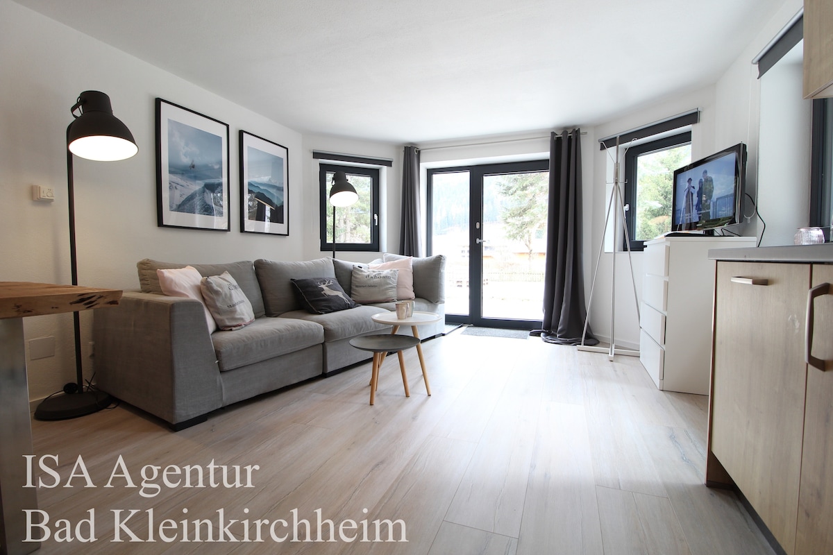 A cozy living area features a light gray sofa facing a wall-mounted TV, complemented by a small side table. Natural light enters through large glass doors that open to a terrace. Tasteful black-and-white artwork adorns the walls, enhancing the calming atmosphere.