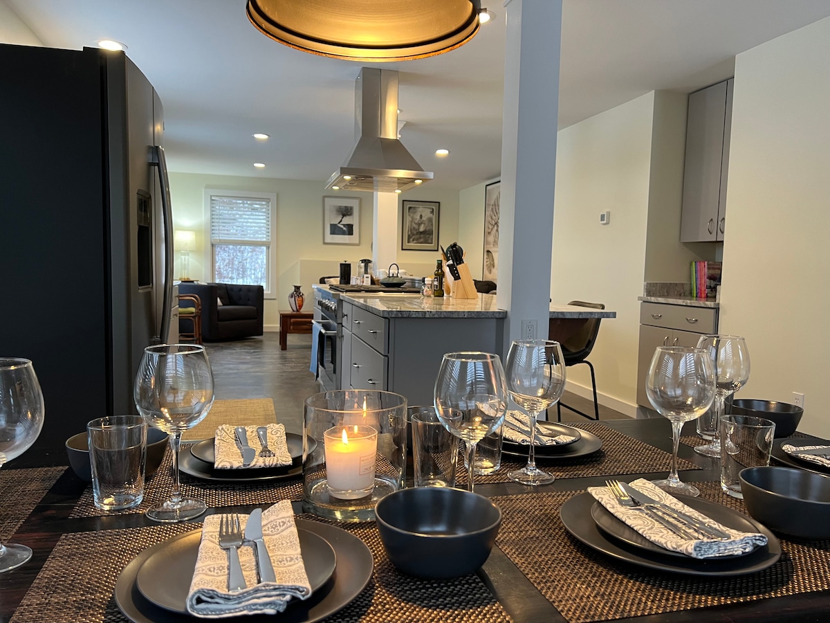 An elegantly set dining table displays black plates, clear glassware, and cloth napkins. A lit candle adds a soft glow, while a modern kitchen is visible in the background, featuring stainless steel appliances and a cozy living area with seating.