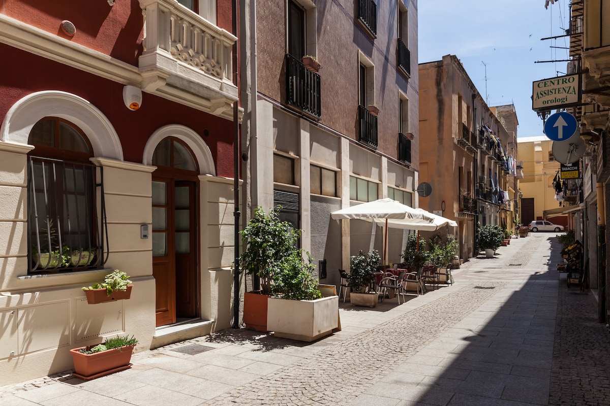A charming street view showcases a well-maintained road lined with colorful buildings. Outdoor seating is visible under a white canopy, with potted plants adding a touch of greenery. The scene reflects the vibrant atmosphere of a pedestrian-friendly area.