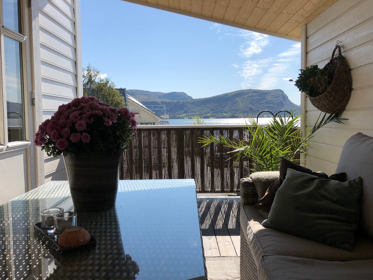 A seating area on a covered terrace is displayed, featuring a glass table and a comfortable sofa adorned with cushions. A potted plant and flowers are positioned on the table, offering a view of the surrounding mountains and blue sky in the background.
