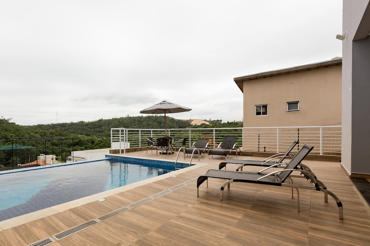An outdoor area features a modern infinity pool beside a spacious wooden deck. Several lounge chairs are positioned for relaxation, and a large umbrella provides shade. A dining table is visible in the background, surrounded by greenery, creating a serene atmosphere.