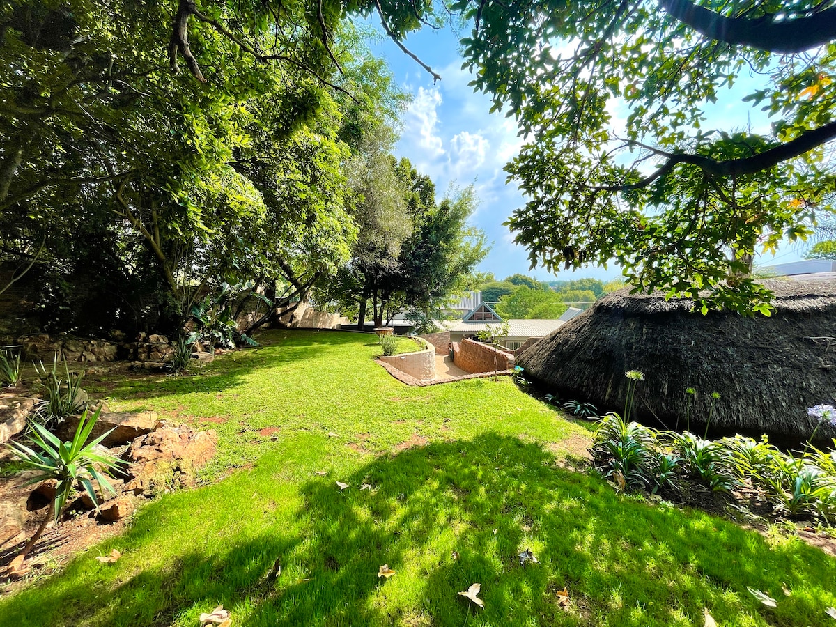 A spacious garden is shown, featuring lush green grass and various plants surrounded by trees. A small pool is visible in the background, alongside a traditional thatched-roof structure, creating a relaxing outdoor environment for guests.