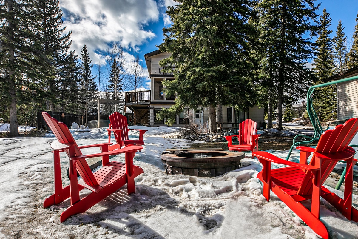 A serene outdoor space features a fire pit surrounded by four red Adirondack chairs, set against a backdrop of snow and trees. The two-story chalet in the distance offers a spacious deck, integrating seamlessly with the natural surroundings.
