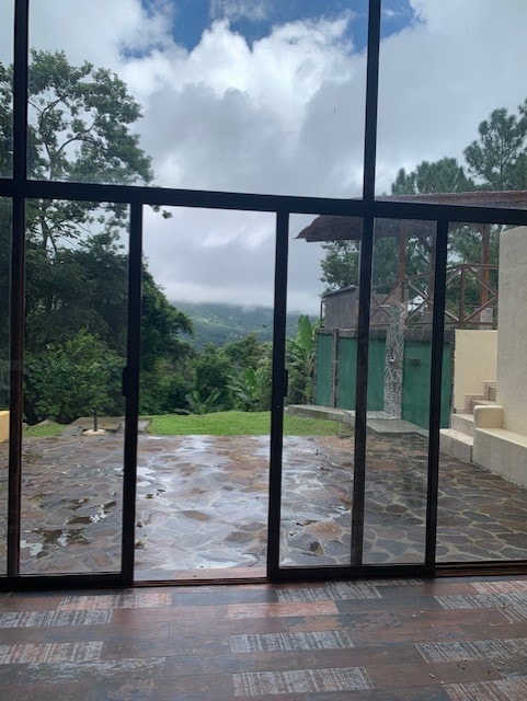 A large glass sliding door provides a view of the lush green landscape outside, framed by tall foliage and a cloudy sky. The stone patio below glistens with moisture, indicating recent rain, while a tranquil atmosphere is felt throughout the space.