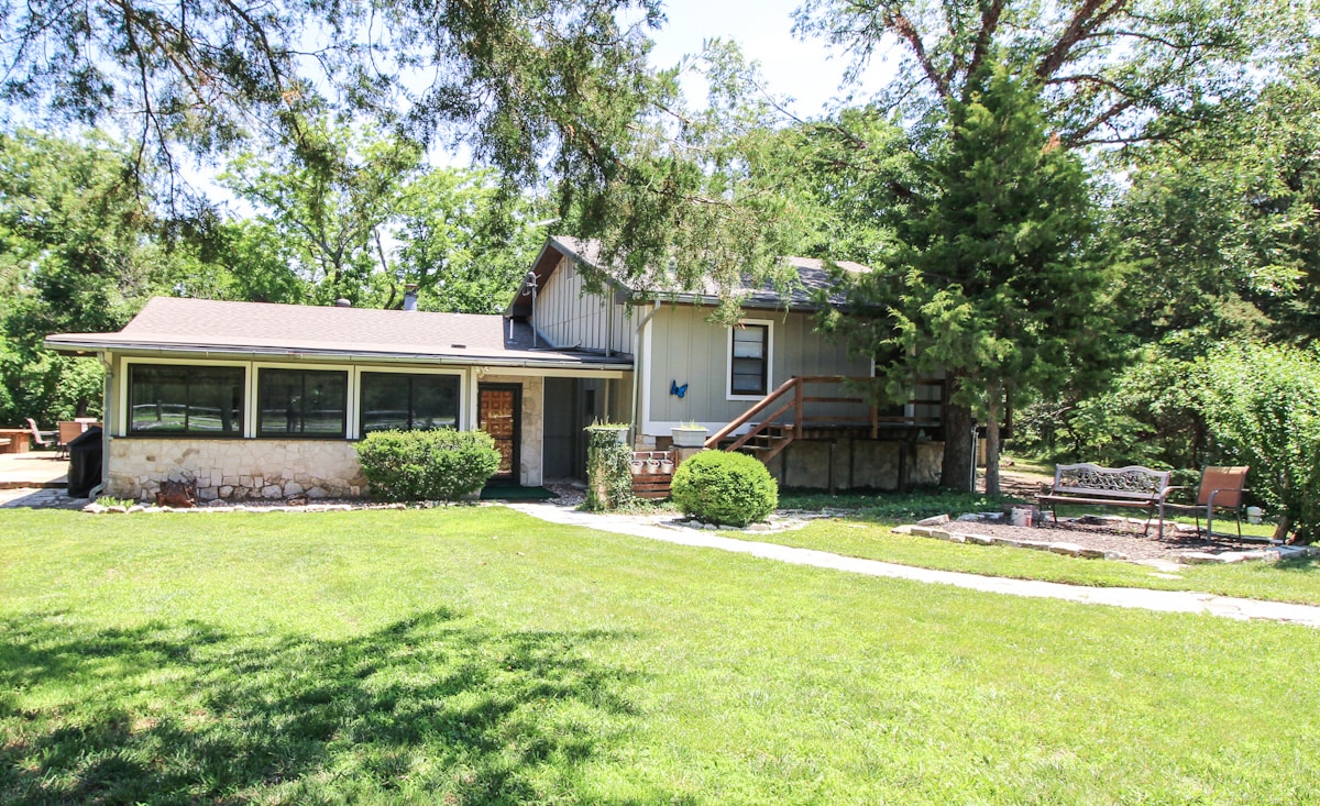 The exterior of a spacious home is showcased, featuring a well-manicured lawn and stone pathways. The structure combines a modern addition with a traditional cabin aesthetic, surrounded by trees that provide a sense of privacy. A cozy seating area is visible in the garden space.