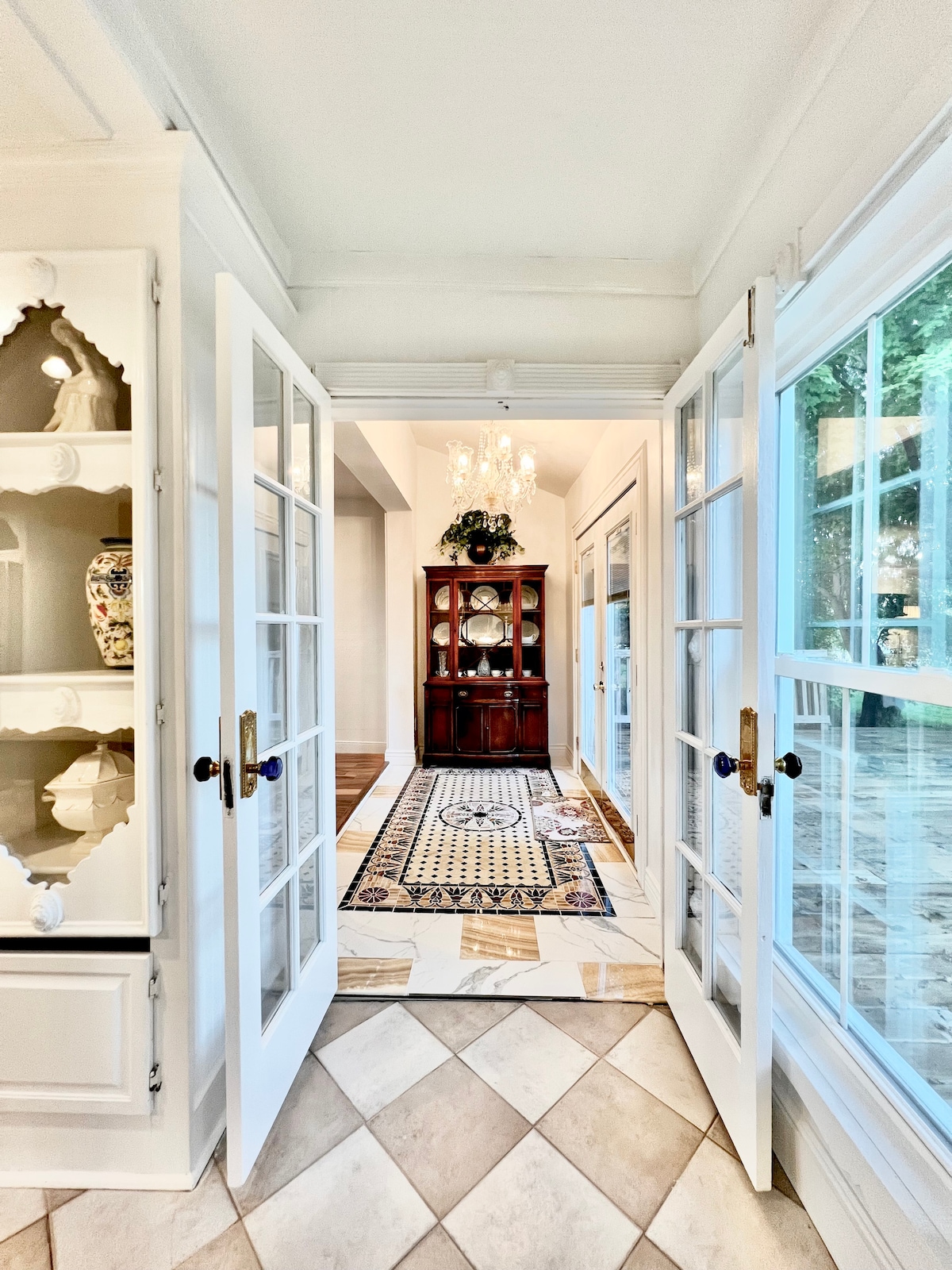 A bright entrance is framed by French doors, leading to a decorative hallway. A classic chandelier hangs above, illuminating the patterned tile floor. An elegant wooden cabinet showcases decorative items, while large windows bring in natural light, enhancing the airy atmosphere.