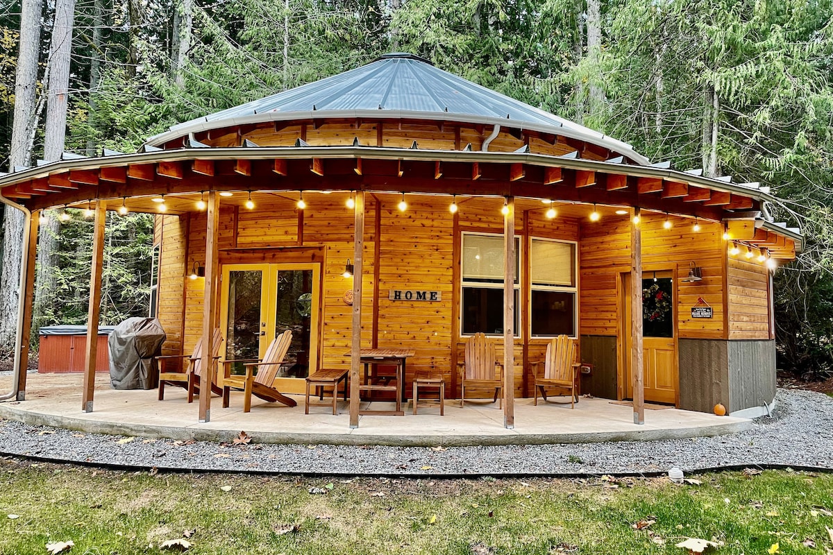 The exterior of a roundhouse is displayed, featuring a wooden facade surrounded by tall trees. A covered porch with several wooden chairs invites relaxation, while soft lighting enhances the welcoming atmosphere. The gravel path leads to the entrance, offering a serene connection to nature.