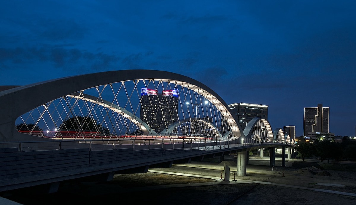 A modern pedestrian bridge is illuminated against a twilight sky, showcasing distinct arched structures and geometric patterns. The backdrop features a skyline of city buildings, with soft lighting highlighting their contours, creating a serene urban ambiance.