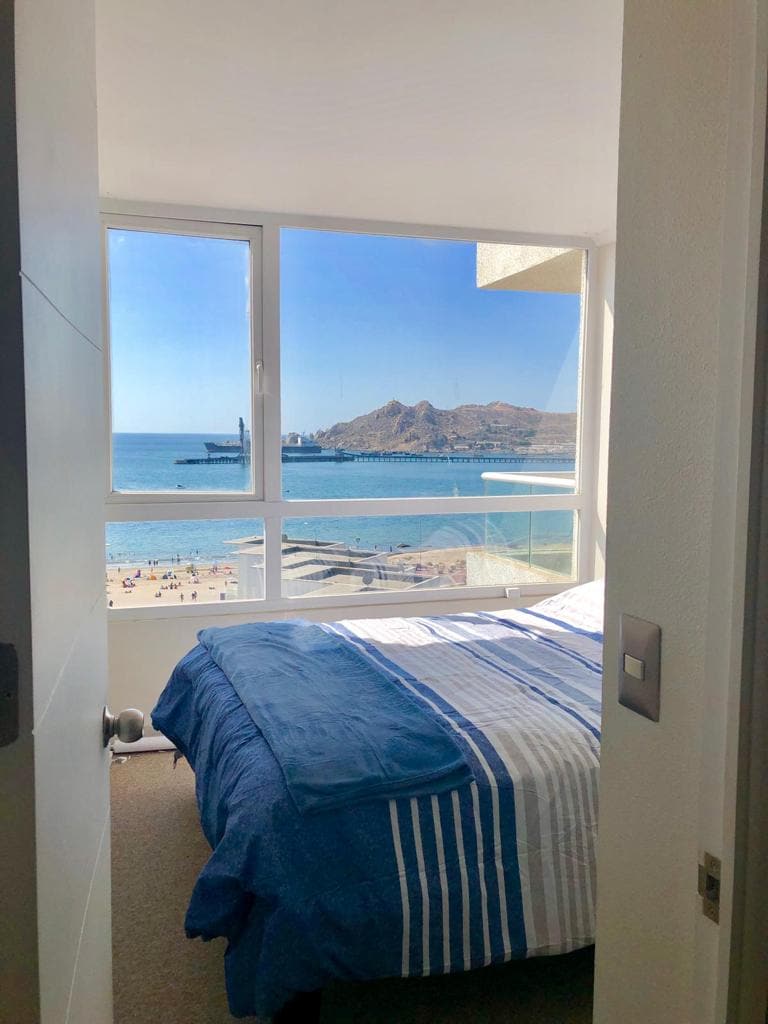 A bedroom is seen through an open door, featuring a spacious bed adorned with blue and white striped bedding. Large windows provide a view of the beach and ocean, allowing natural light to brighten the room.
