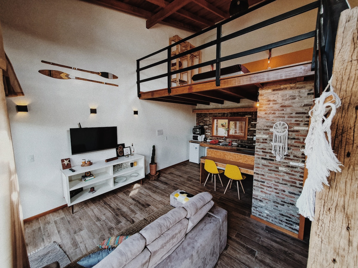 A spacious living area features a comfortable sofa in neutral tones and a flat-screen television mounted on the wall. The open kitchen, equipped with modern appliances, includes a wooden bar with yellow chairs. A loft with a wooden railing overlooks the room.