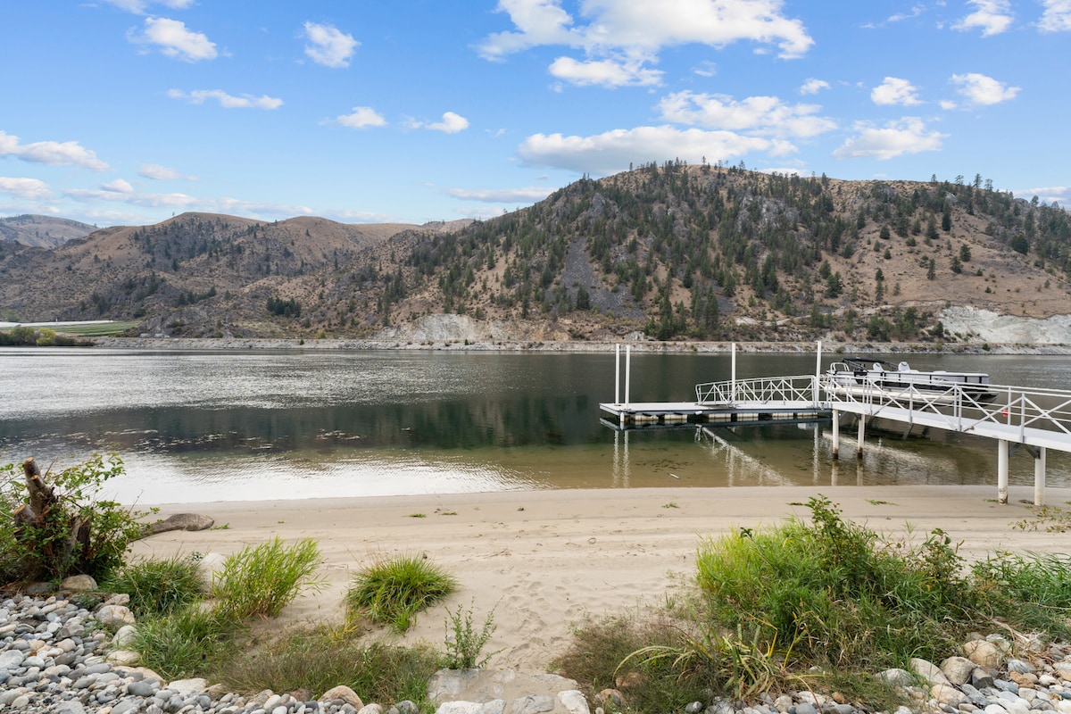 A sandy beach leads to a calm section of the Columbia River, framed by gently rolling hills. A white dock extends into the water, creating a peaceful spot for activities. Lush greenery and rocks are visible along the beach, enhancing the serene natural setting.
