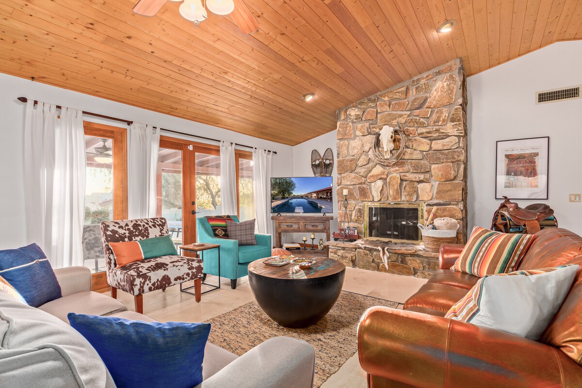 A cozy living room features a mix of seating options, including a leather sofa and colorful armchairs. A stone fireplace adds character, while large windows allow natural light and views of the outdoors. Wooden ceilings and various decor elements enhance the southwestern theme.