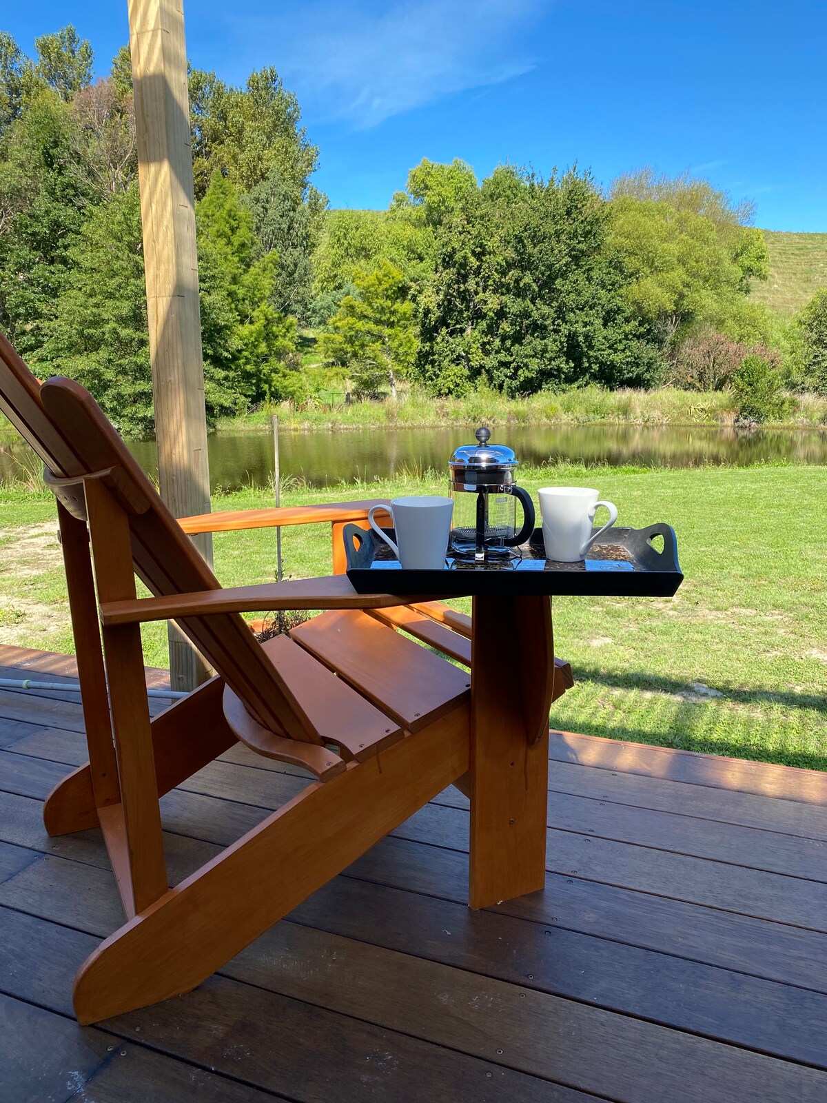 A wooden chair positioned on a deck overlooks a serene pond, showcasing a tray with two cups and a French press. Lush greenery surrounds the area, and a clear blue sky enhances the peaceful setting.