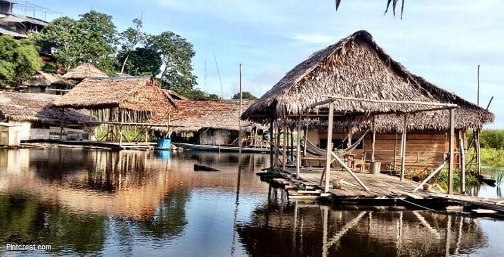 Traditional wooden structures with thatched roofs are situated along the water's edge. A wooden walkway connects the buildings, reflecting on the calm water surface, surrounded by lush greenery and trees.