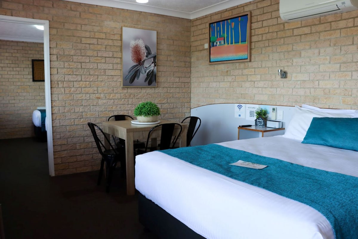 A well-lit room features a spacious bed with crisp white linens and a turquoise accent. A small wooden dining table with black chairs is positioned nearby. Artwork decorates the wall, and air conditioning unit is mounted above, enhancing the room's comfort and functionality.