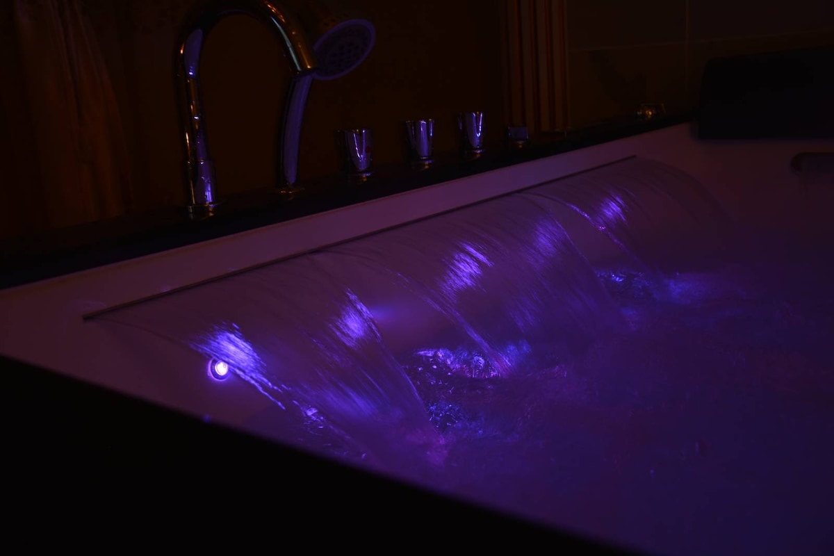 A private jacuzzi is displayed, showcasing gentle water cascades illuminated by soft purple lighting. The tranquil setting highlights the soothing flow of water against a sleek, modern tub design.