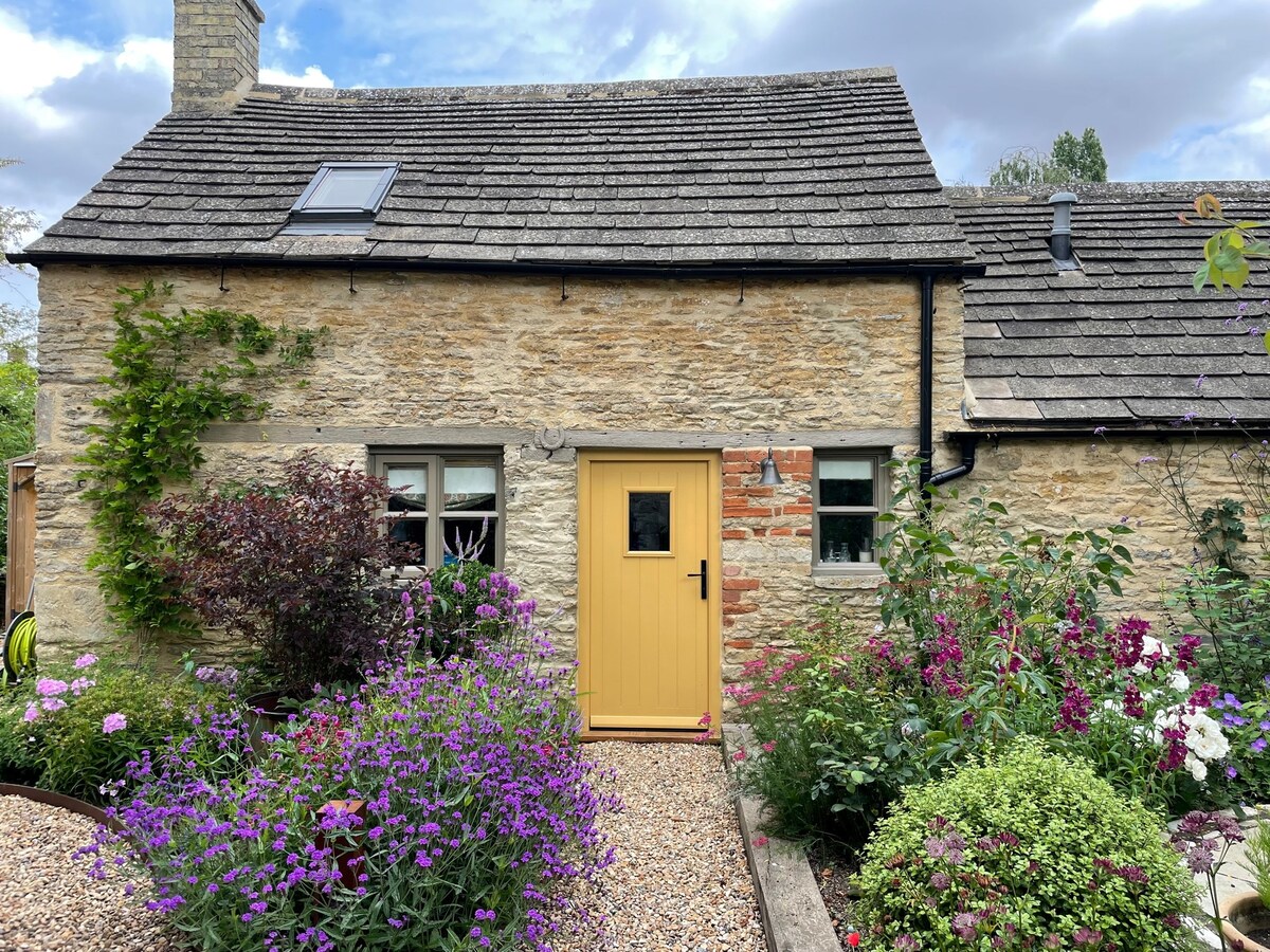 A charming stone cottage features a bright yellow front door, surrounded by colorful flowers and lush greenery. The weathered roof and inviting windows enhance the rustic appeal, while the landscaped pathway draws attention with its pebbles and blooming plants.