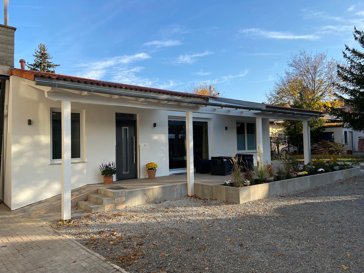 A modern bungalow is depicted with a covered terrace supported by white posts. The entrance features a dark door beside large windows. Outdoor seating is visible on the terrace, surrounded by planters with flowers. A gravel driveway leads up to the home, enhancing its inviting appearance.