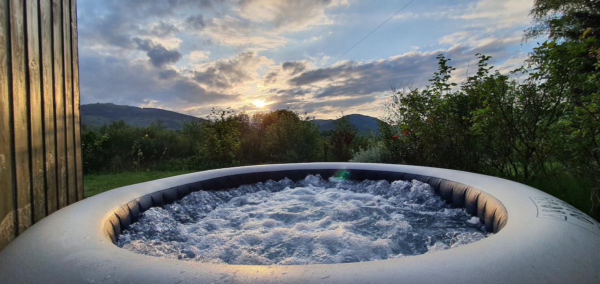 A private jacuzzi is positioned outdoors, surrounded by lush greenery and colorful plants. The water appears inviting as bubbles gently rise to the surface. A scenic landscape of rolling hills is visible in the background under a partly cloudy sky, hinting at a tranquil sunset.