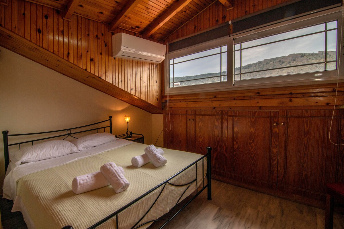 A cozy sleeping area is featured under a wooden ceiling, with a spacious bed dressed in white linens. Two towels are neatly placed at the foot of the bed. Natural light enters through windows framed in wood, highlighting the warm tones of the wooden walls.