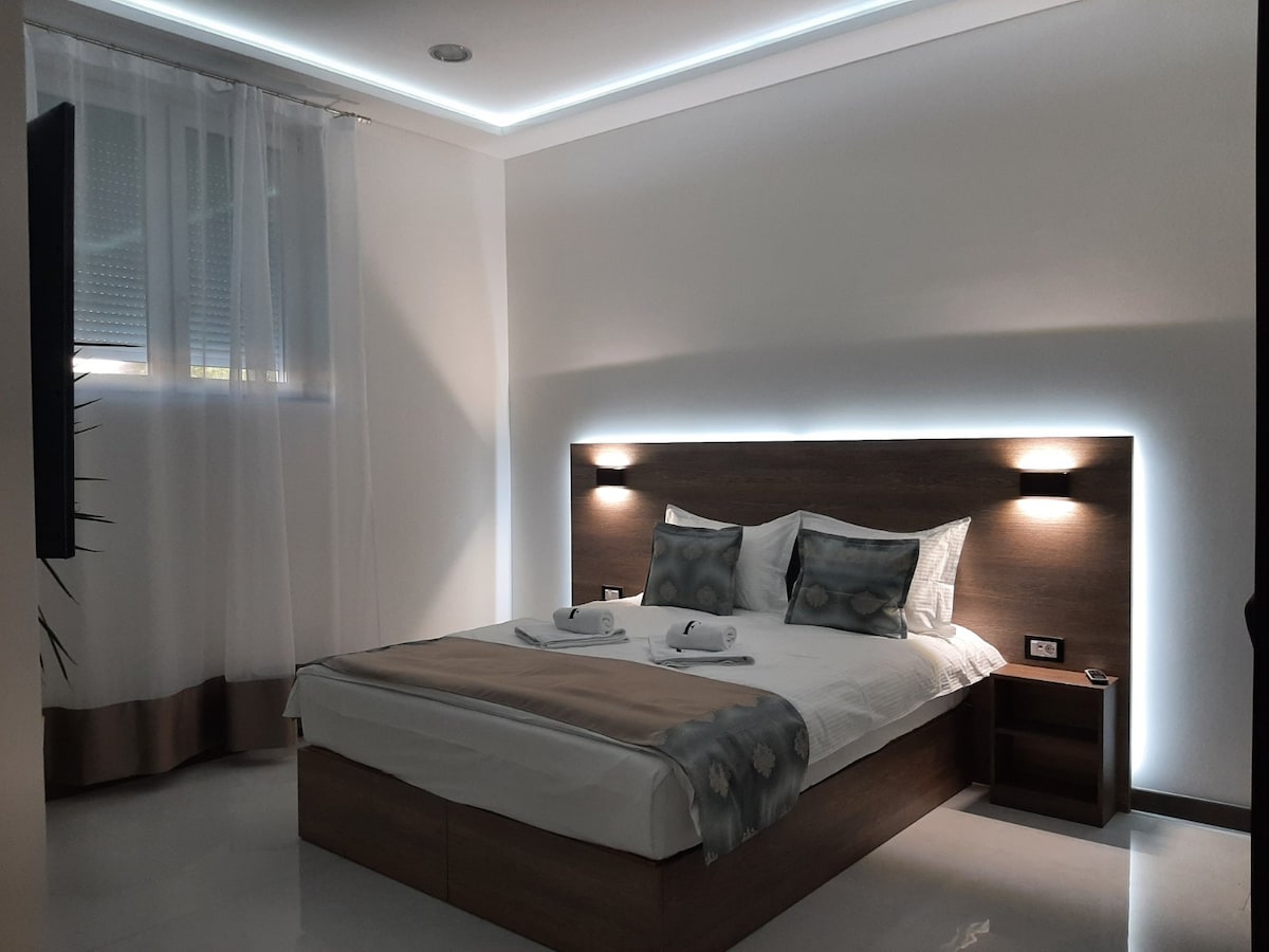 A modern bedroom features a spacious bed with an upholstered headboard, flanked by reading lights. Two rolled towels are arranged neatly on the bed. Soft white curtains diffuse natural light from the window, while subtle lighting along the ceiling adds a calming ambiance.