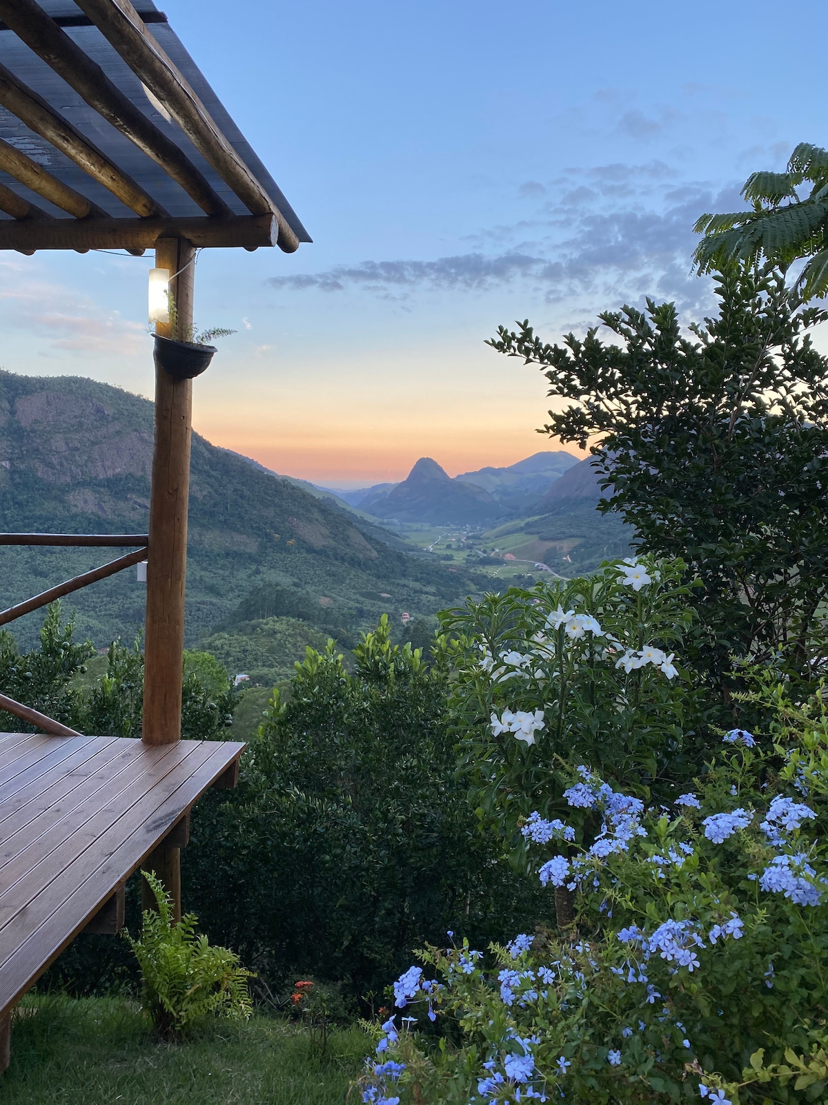 A wooden patio offers a panoramic view of rolling mountains under a soft twilight sky. The landscape features lush greenery and scattered wildflowers, with a calming gradient of colors reflecting the setting sun.