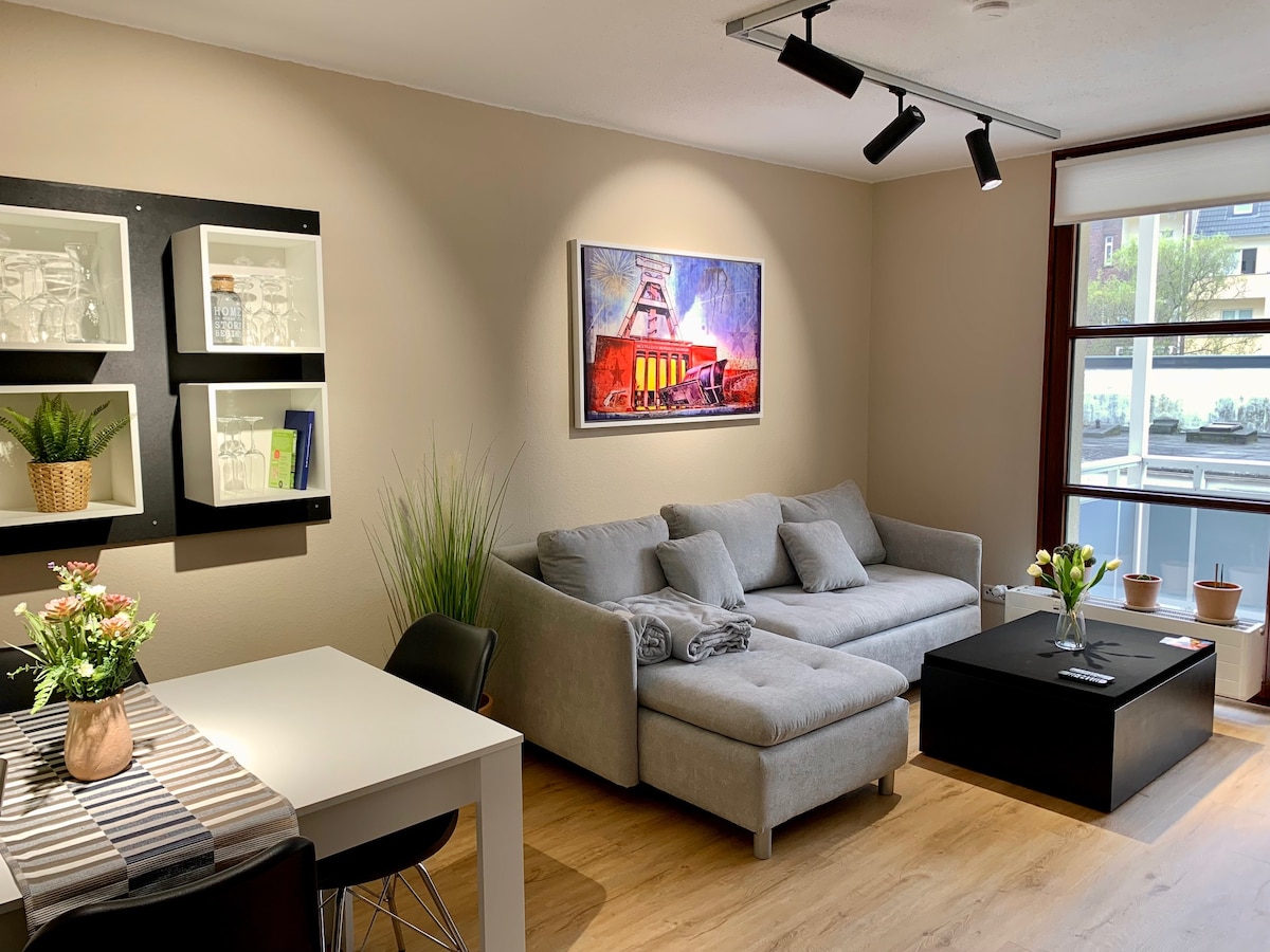 A modern living area features a comfortable gray sectional sofa and a black coffee table. A white dining table is adorned with a striped table runner and a vase of flowers. Artwork depicting a colorful building is displayed on the wall, alongside shelves showcasing decorative items.