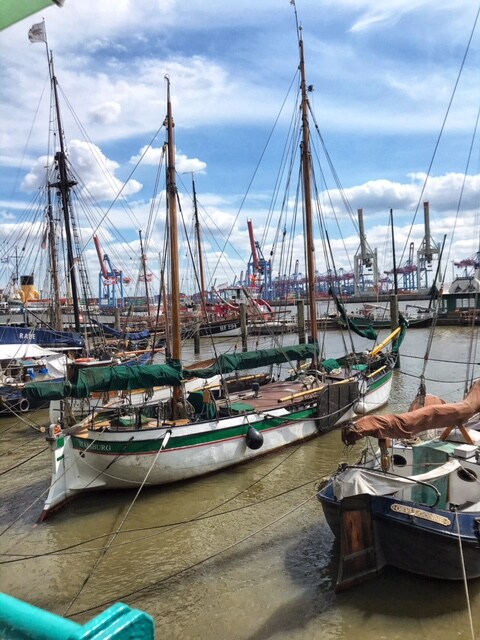 A collection of traditional sailing boats rests at the harbor, with masts reaching towards a partly cloudy sky. Background cranes and industrial structures indicate a working port. Calm waters reflect the boats, creating a peaceful scene ideal for maritime enthusiasts.